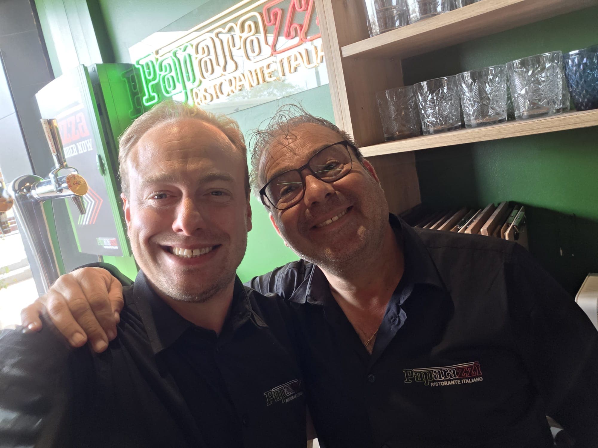 Dos hombres sonrientes con uniformes de Paparazzi Ristorante Italiano posan frente a una barra verde.